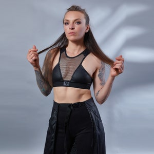 May include: A woman wearing a black mesh and leather sports bra with a black belt and trousers. She has long dark hair and tattoos on her arms.