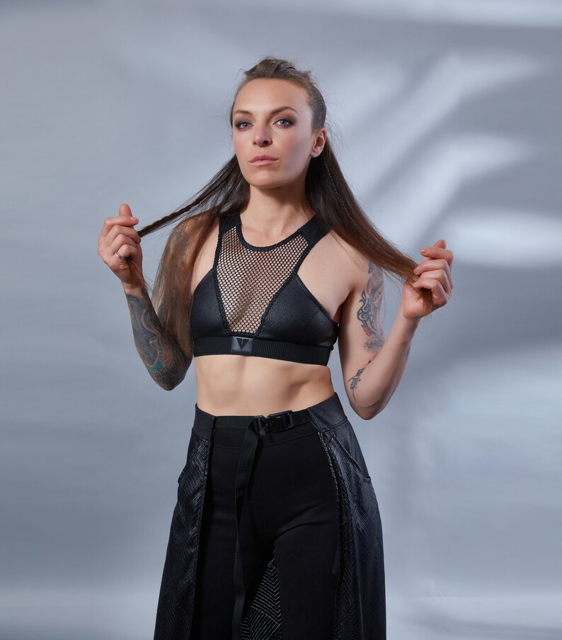 May include: A woman wearing a black mesh and leather sports bra with a black belt and trousers. She has long dark hair and tattoos on her arms.