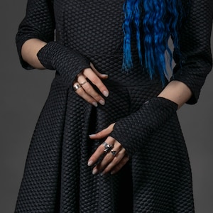 May include: A black dress with a textured, quilted-style pattern featuring a fitted bodice and a flared skirt. The garment includes matching black fingerless gloves made from the same quilted fabric, worn on the forearms and hands.