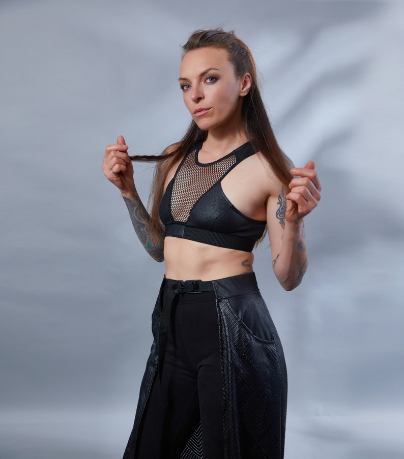 May include: A woman wearing a black mesh and leather sports bra with a black skirt and trousers. She has tattoos on her arms.