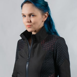 May include: A woman with blue hair wearing a black jacket with a red and black honeycomb pattern. The jacket has a zipper and a high collar.