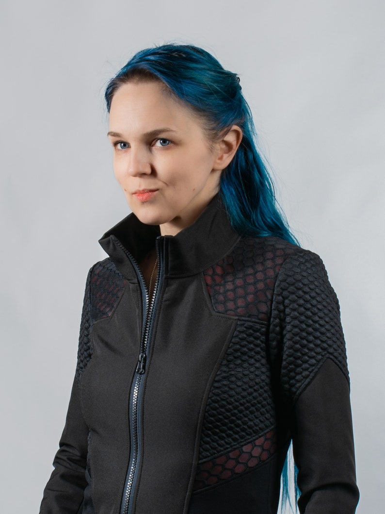 May include: A woman with blue hair wearing a black jacket with a red and black honeycomb pattern. The jacket has a zipper and a high collar.