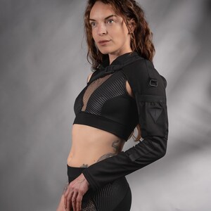K&ouml;nnte beinhalten: Eine Frau tr&auml;gt ein schwarzes Crop-Top mit einem Mesh-Einsatz und ein schwarzes Langarmshirt mit einem Schulterriemen. Das Shirt hat einen dreieckigen Patch am Oberarm. Sie tr&auml;gt eine schwarze Leggings mit einem Mesh-Einsatz an der Seite.