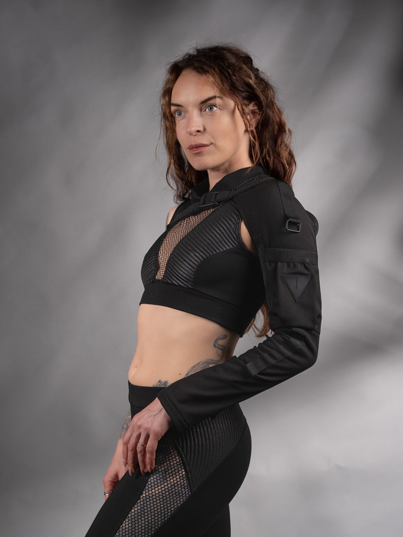 K&ouml;nnte beinhalten: Eine Frau tr&auml;gt ein schwarzes Crop-Top mit einem Mesh-Einsatz und ein schwarzes Langarmshirt mit einem Schulterriemen. Das Shirt hat einen dreieckigen Patch am Oberarm. Sie tr&auml;gt eine schwarze Leggings mit einem Mesh-Einsatz an der Seite.