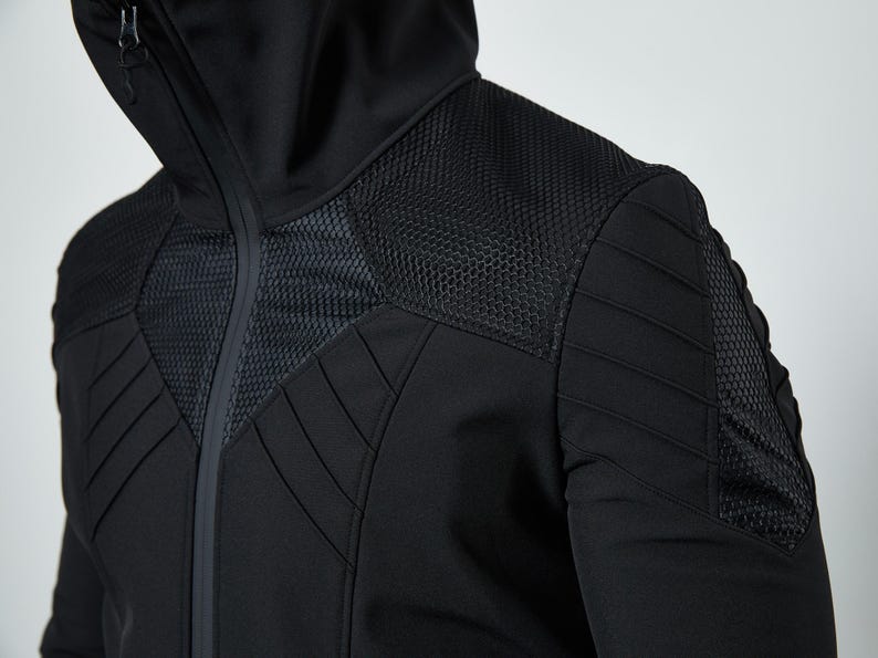 May include: A black hooded jacket featuring a high collar, a full-length front zipper, and textured mesh panels on the chest and shoulders. The garment includes padded, stitched detailing across the upper torso and sleeves for a structured look.