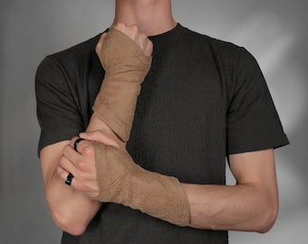 May include: A pair of tan, textured leather fingerless wrist wraps that cover the lower forearm and the back of the hand, featuring a thumb hole and a speckled surface pattern. The wraps are worn on both wrists, extending from the knuckles to the mid-forearm.