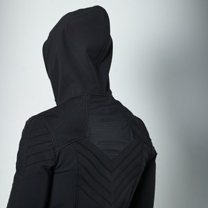 May include: A black hooded jacket featuring a textured mesh panel across the upper back and shoulders, complemented by horizontal and chevron-shaped stitched detailing that creates a structured, layered appearance across the rear of the garment.