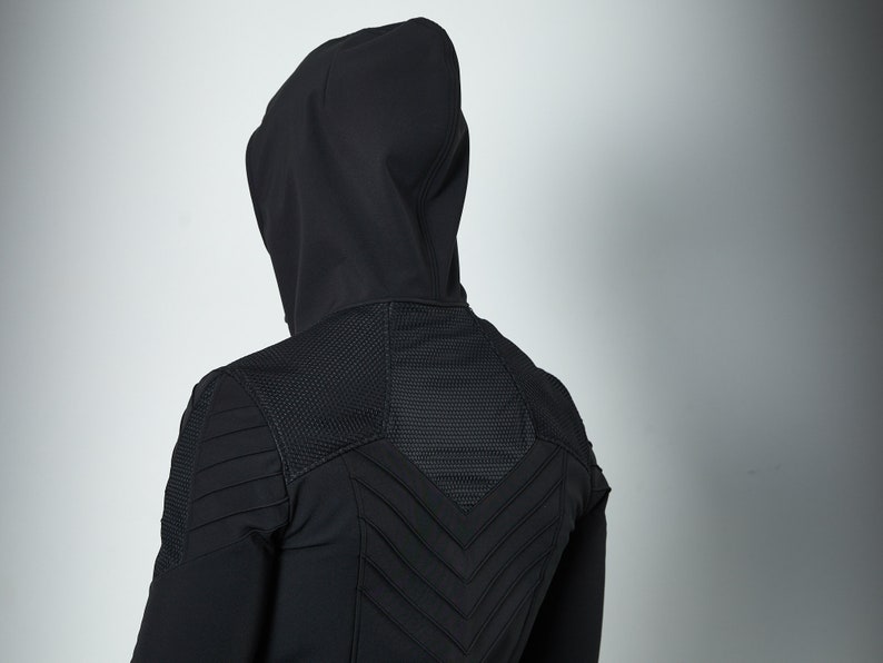 May include: A black hooded jacket featuring a textured mesh panel across the upper back and shoulders, complemented by horizontal and chevron-shaped stitched detailing that creates a structured, layered appearance across the rear of the garment.