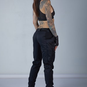 Puede incluir: Una mujer con pantalones negros y un sujetador deportivo negro con espalda de malla. Tiene tatuajes en los brazos y la espalda. Lleva guantes negros y zapatos negros.