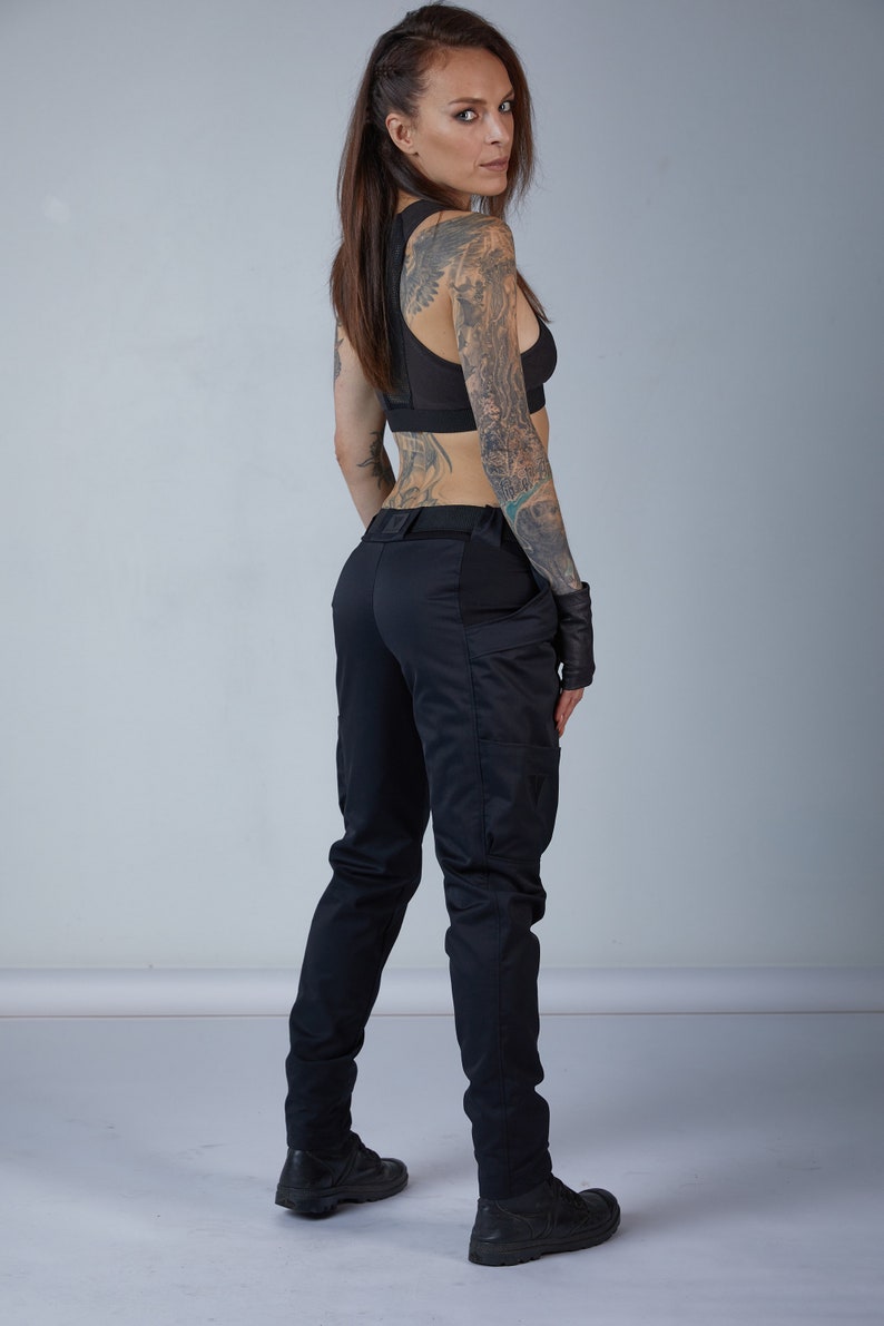 Puede incluir: Una mujer con pantalones negros y un sujetador deportivo negro con espalda de malla. Tiene tatuajes en los brazos y la espalda. Lleva guantes negros y zapatos negros.