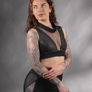 K&ouml;nnte beinhalten: Eine Frau tr&auml;gt ein schwarzes Crop-Top mit einem Mesh-Einsatz und schwarze Leggings mit Mesh-Eins&auml;tzen. Sie hat Tattoos auf ihren Armen.