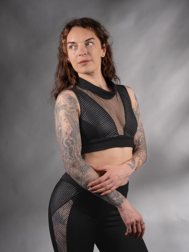 K&ouml;nnte beinhalten: Eine Frau tr&auml;gt ein schwarzes Crop-Top mit einem Mesh-Einsatz und schwarze Leggings mit Mesh-Eins&auml;tzen. Sie hat Tattoos auf ihren Armen.
