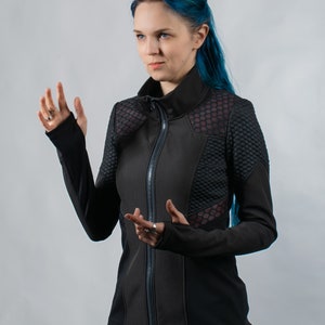 May include: A woman with blue hair is wearing a black zippered jacket with a geometric pattern. The jacket has a high collar and long sleeves.