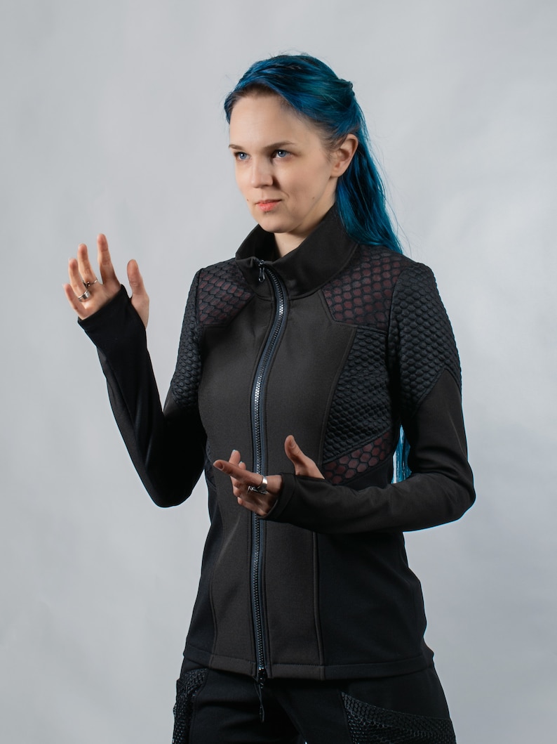 May include: A woman with blue hair is wearing a black zippered jacket with a geometric pattern. The jacket has a high collar and long sleeves.