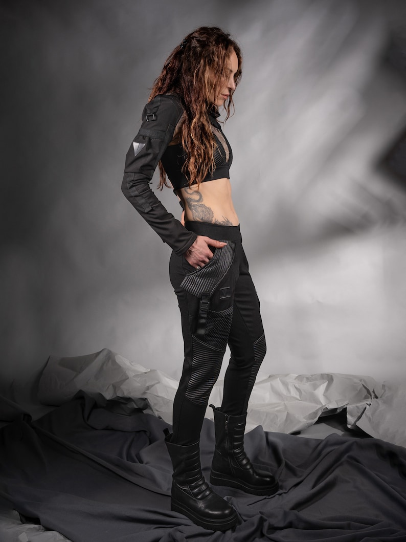 K&ouml;nnte beinhalten: Eine Frau tr&auml;gt ein schwarzes, lang&auml;rmliges Crop-Top mit einem Mesh-Einsatz und eine schwarze Hose mit Rei&szlig;verschlussdetail. Sie tr&auml;gt schwarze Stiefel und hat lange braune Haare.