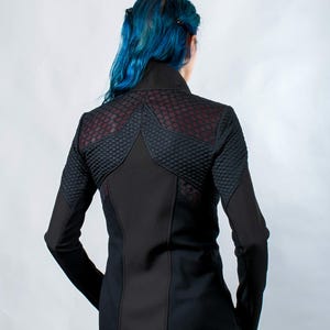 May include: A black long-sleeved jacket featuring a high collar and a textured, scale-like pattern across the upper back and shoulders. The garment includes solid black panels along the sides and sleeves, creating a structured, fitted appearance.