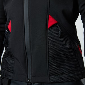 May include: A black jacket featuring a central vertical zipper, two large black mesh pockets with zipper closures, and vibrant red fabric accents positioned above each pocket. The garment is constructed from a smooth, solid black material.