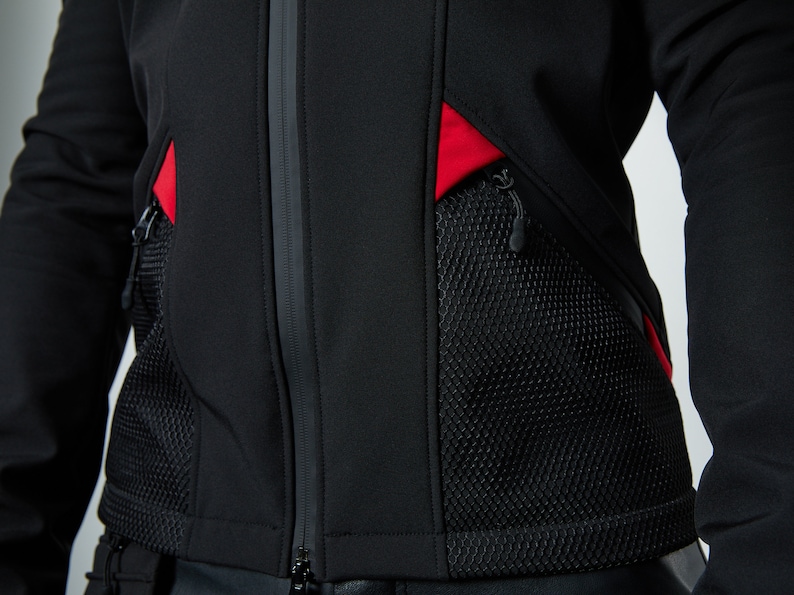 May include: A black jacket featuring a central vertical zipper, two large black mesh pockets with zipper closures, and vibrant red fabric accents positioned above each pocket. The garment is constructed from a smooth, solid black material.