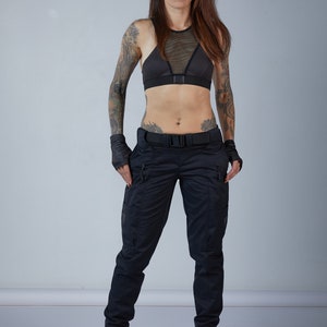 Puede incluir: Una mujer con un sujetador deportivo negro con un panel de malla y pantalones cargo negros. Lleva guantes negros sin dedos y tiene tatuajes en los brazos.