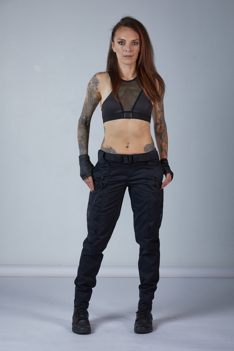 Puede incluir: Una mujer con un sujetador deportivo negro con un panel de malla y pantalones cargo negros. Lleva guantes negros sin dedos y tiene tatuajes en los brazos.