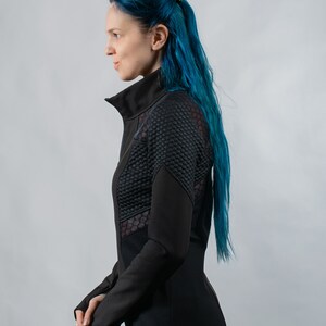 May include: A black zippered jacket with a mesh panel on the side. The jacket has a stand-up collar and long sleeves. The model has blue hair.