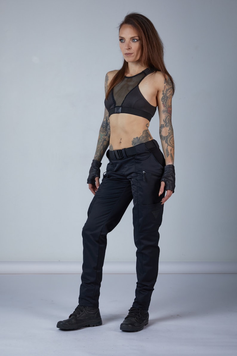Puede incluir: Una mujer con un sujetador deportivo de malla negra y pantalones cargo negros con un cintur&oacute;n negro. Lleva guantes negros sin dedos y tiene tatuajes en los brazos.