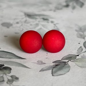 Könnte beinhalten: Zwei leuchtend rote, kugelförmige Ohrstecker. Die Ohrringe sind nebeneinander vor einem Hintergrund aus weißem und grauem Blumenmuster platziert. Die Ohrringe haben eine matte Oberfläche und scheinen aus einem festen Material zu bestehen.