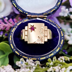 Op de afbeelding: Een gouden zegelring met een rode edelsteen die in het midden van een rechthoekige bovenkant is gezet. De ring zit in een blauwe fluwelen ringdoos.