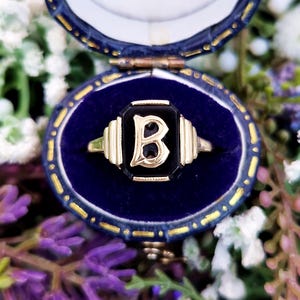 May include: A gold ring with a black onyx center featuring the letter "B" in a serif font. The ring is set in a blue velvet jewelry box with a gold trim. The background includes purple and white flowers.