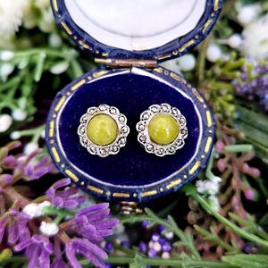 Könnte beinhalten: Vintage Ohrstecker mit einem hellgrünen Mittelstein, gefasst in silberfarbenem Metall mit floralem Design. Die Ohrringe werden in einer offenen, blauen Samt-Schmuckschatulle mit goldenem Rand präsentiert, umgeben von Grün.