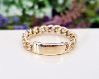 May include: A gold-colored ring with a curb chain design and a rectangular plate. The ring is set against a white surface with blurred floral background. The ring's design is a classic and elegant style.