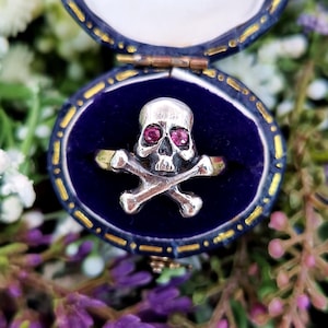 May include: A silver skull ring with red gemstone eyes and crossed bones. The ring is displayed in a blue velvet jewelry box with a gold trim, surrounded by flowers. The ring is a unique piece of jewelry.