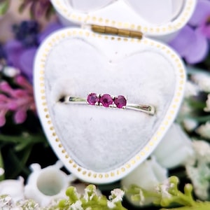 以下が含まれることがあります： 3つの丸い赤い宝石が並んだシルバーリング。 リングは、金色の留め金が付いたハート型の白いジュエリーボックスに入れられ、花に囲まれて展示されています。