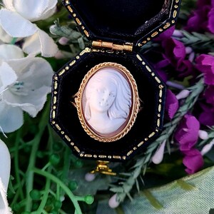 Op de afbeelding: Een cameo ring met een witte cameo van een vrouwen gezicht, gezet in een gouden ovale lijst. De ring zit in een zwarte ringdoos met een gouden rand.