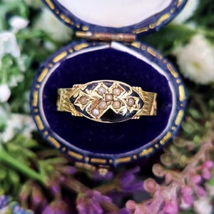 Puede incluir: Un anillo de oro con un centro de esmalte negro, adornado con pequeñas perlas en un patrón floral. El anillo se presenta en un joyero de terciopelo azul, rodeado de flores. El diseño del anillo es intrincado y detallado.