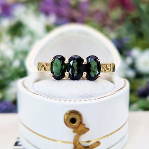 Puede incluir: Un anillo de oro con tres piedras preciosas ovaladas de color verde oscuro. El anillo está engastado en una banda de oro con filigrana decorativa en los laterales. Se muestra en una caja de joyería blanca con ribetes dorados.