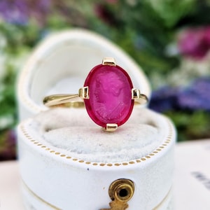 May include: A gold ring with a large, oval, magenta-coloured stone. The stone features a carved profile. The ring is set in a white jewellery box with gold accents. The background is a soft focus of flowers.