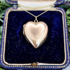 Könnte beinhalten: Ein herzförmiger Medaillon-Anhänger aus Gold an einer dünnen Goldkette. Das Medaillon hat eine polierte Oberfläche mit zarten, eingravierten blattartigen Mustern an den Seiten. Es wird in einer quadratischen, mit marineblauem Samt ausgekleideten Schmuckschatulle präsentiert.