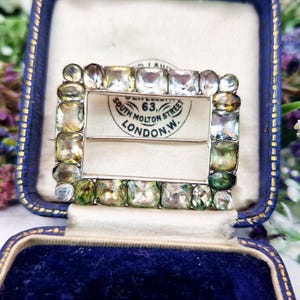 May include: A rectangular brooch featuring a border of multi-colored, faceted stones in shades of green, yellow, and clear, set in a silver-toned metal frame. The brooch is displayed inside a dark blue velvet-lined jewelry box with text.
