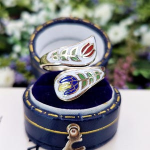 May include: A gold-toned ring with a floral design, featuring red and blue enamel flowers with green leaves. The ring is displayed in a blue jewelry box with a gold clasp, set against a blurred floral background.