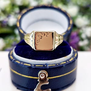 May include: A gold signet ring with a rectangular face, featuring an engraved design. The ring is displayed inside a blue velvet-lined jewellery box. The ring's band has a detailed design on the sides.