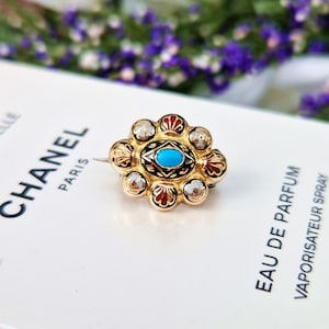 May include: A gold-toned brooch with a turquoise center stone surrounded by a black and white enamel design. The brooch is set with small white pearls around the outer edge.