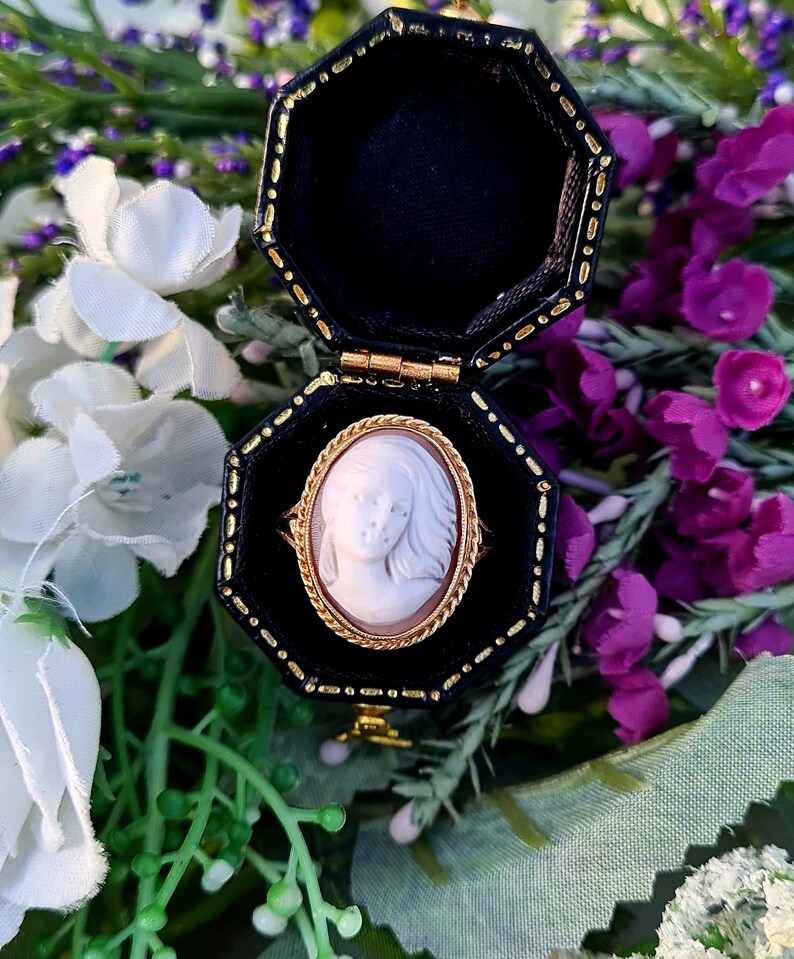 Op de afbeelding: Een cameo ring met een witte cameo van een vrouwen gezicht, gezet in een gouden ovale lijst. De ring zit in een zwarte ringdoos met een gouden rand.