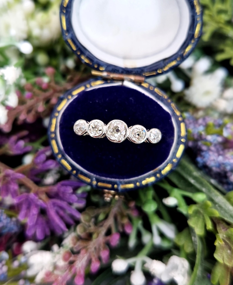 May include: A vintage bar pin with five round diamonds set in a row, displayed in a blue velvet jewelry box. The diamonds sparkle against the dark blue background. The box is open, revealing the pin. The background is a soft focus of flowers.