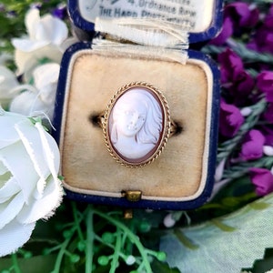 Op de afbeelding: Een gouden ring met een cameo van een vrouwen gezicht. De cameo is gezet in een gouden ovale lijst met een touwontwerp. De ring bevindt zich in een blauwe fluwelen ringdoos.