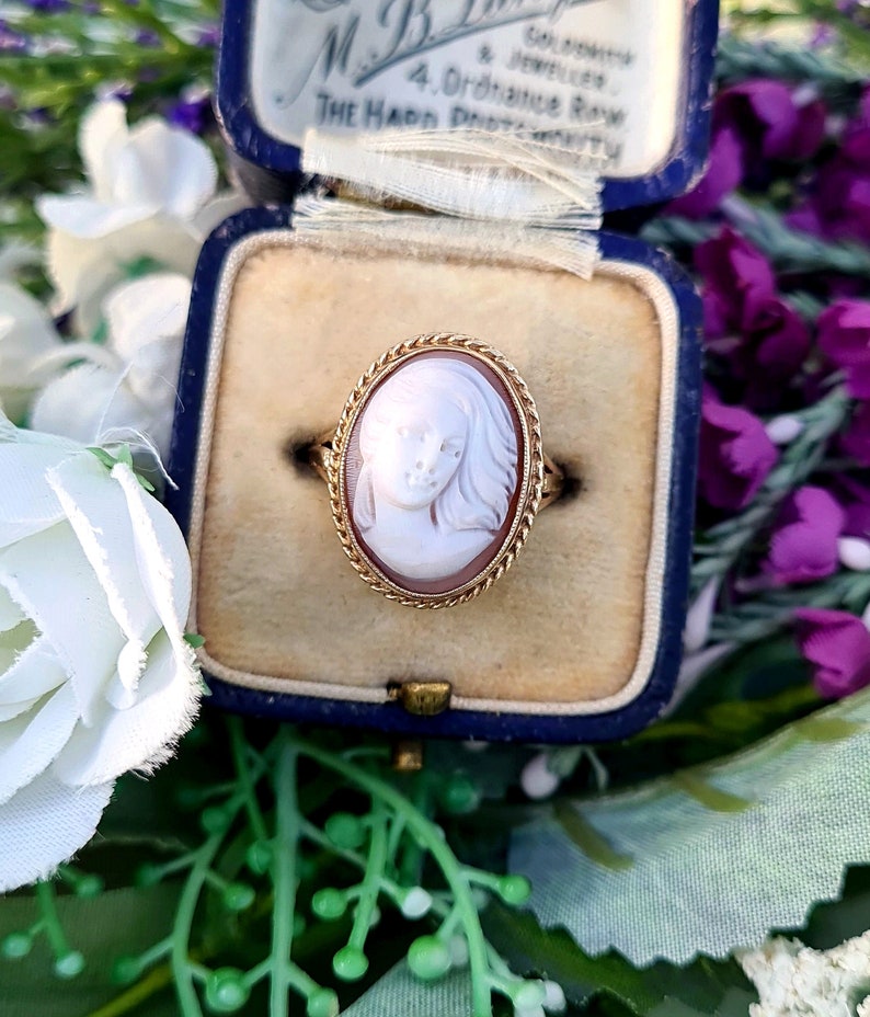 Op de afbeelding: Een gouden ring met een cameo van een vrouwen gezicht. De cameo is gezet in een gouden ovale lijst met een touwontwerp. De ring bevindt zich in een blauwe fluwelen ringdoos.