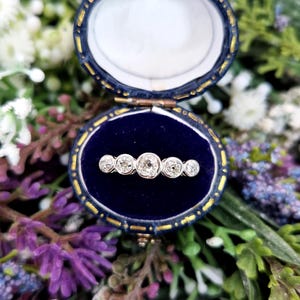 May include: An antique, five-stone diamond ring displayed in a blue velvet jewelry box. The ring features five round-cut diamonds set in a row. The box is oval-shaped with gold trim and is open, revealing the ring against a dark blue interior.
