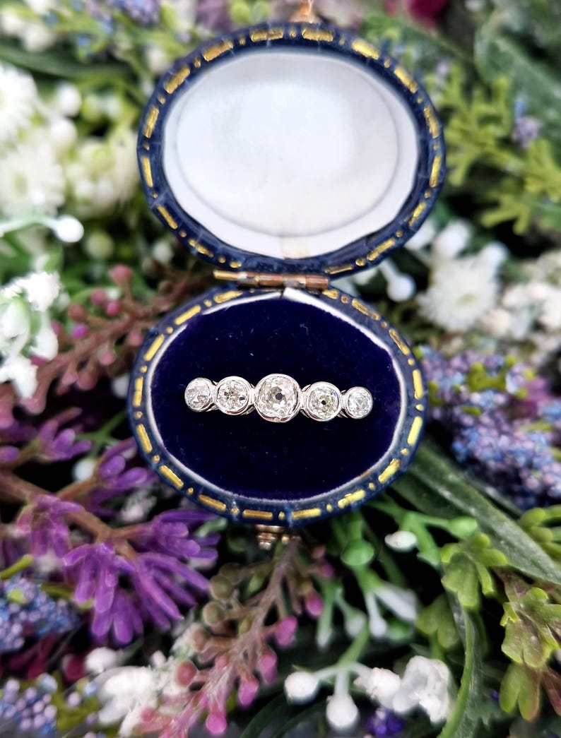 May include: An antique, five-stone diamond ring displayed in a blue velvet jewelry box. The ring features five round-cut diamonds set in a row. The box is oval-shaped with gold trim and is open, revealing the ring against a dark blue interior.