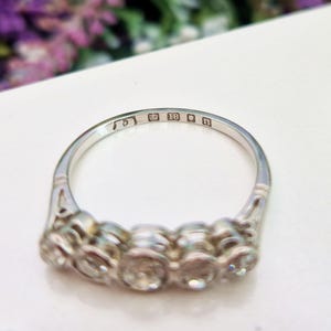 May include: A silver ring with five clear gemstones. The ring has an intricate design on the band and is marked with the number 18 and the letter n. The ring is set on a white surface.