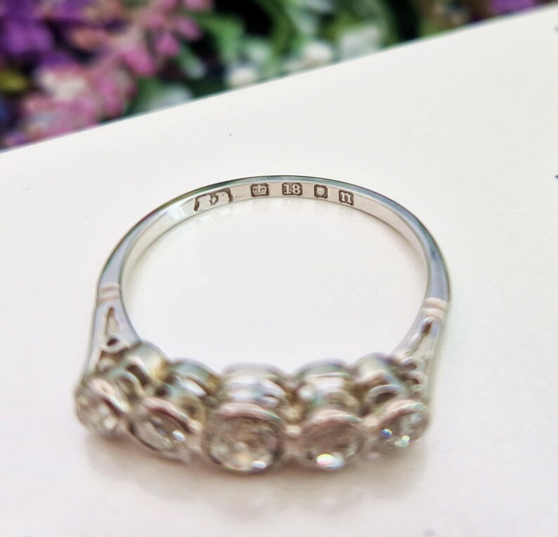May include: A silver ring with five clear gemstones. The ring has an intricate design on the band and is marked with the number 18 and the letter n. The ring is set on a white surface.